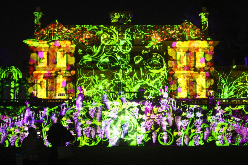 Mansion at night illuminated with green, purple, yellow, and orange lights.