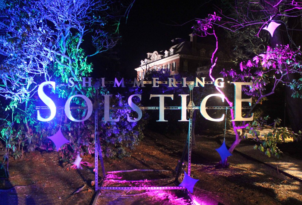 Sign lit up at night in blue, green, and purple hues. Text reads "Shimmering Solstice."